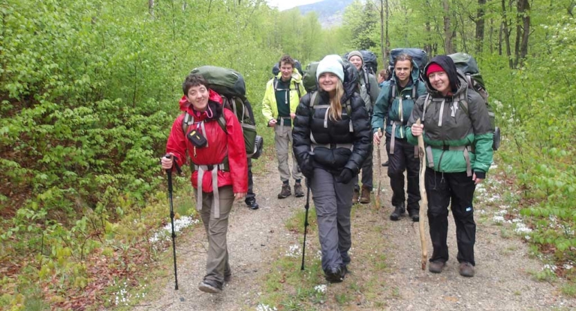 A group of people wearing backpacks and using trekking poles smile for a photo in a wooded area. 